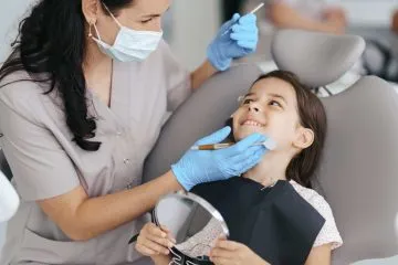 Dentist in mask and gloves examining a smiling young girl holding a mirror