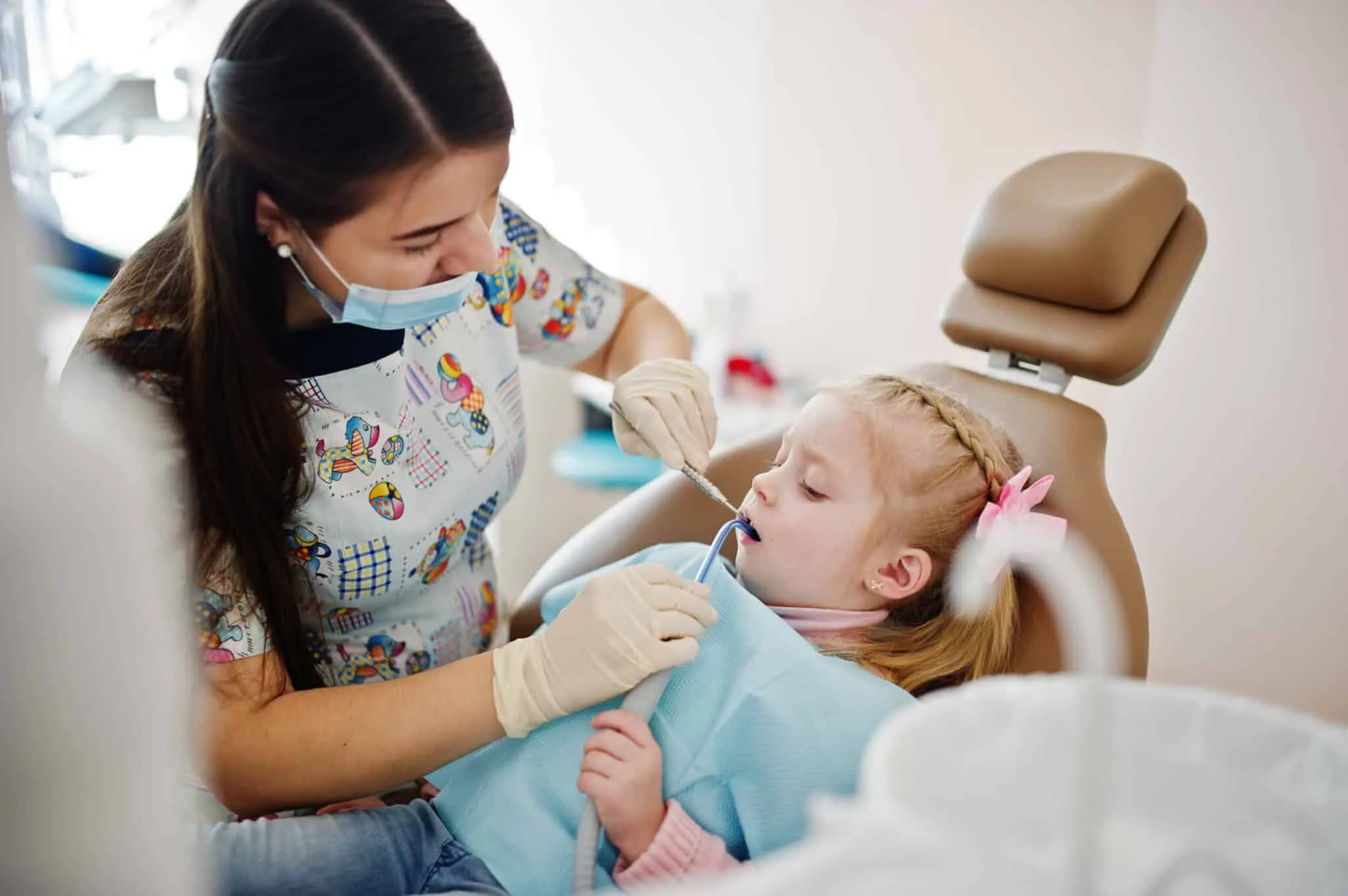Dentist in patterned scrubs examining a young girl’s teeth in a dental chair