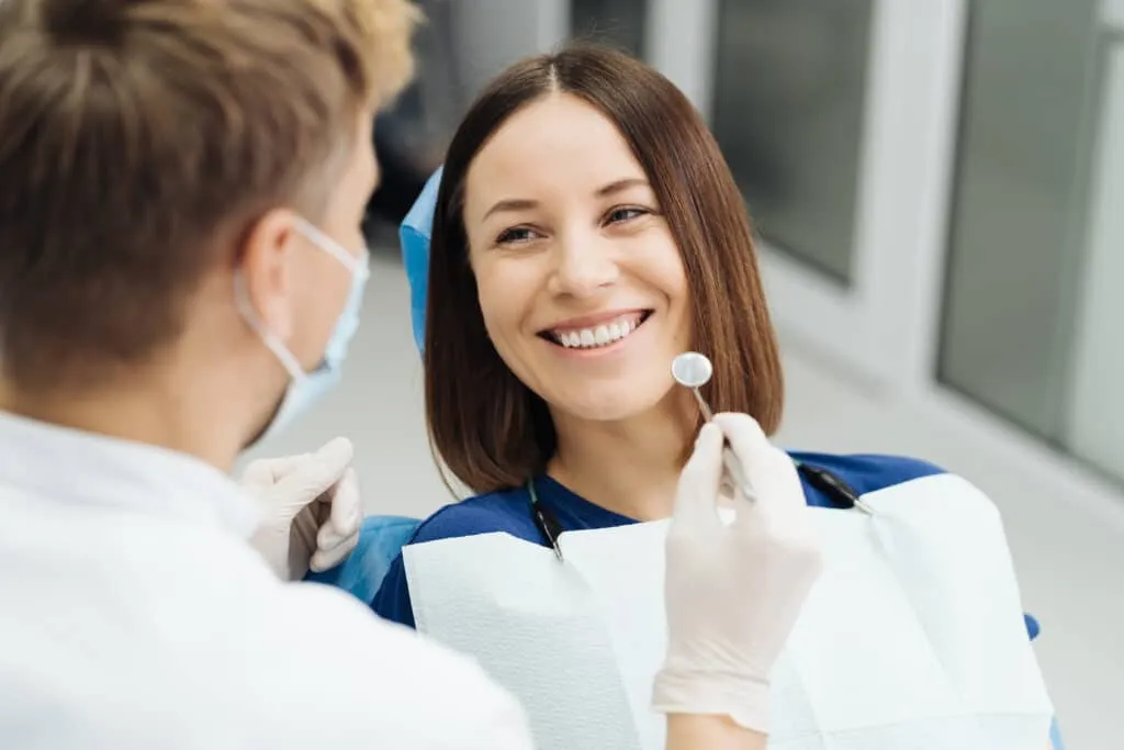Smiling woman in a dental chair being examined by a dentist with a dental mirror.