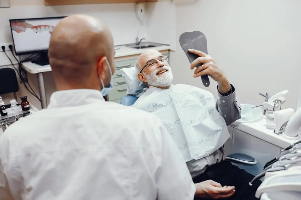 Elderly man in a dental chair smiling while holding a mirror, with a dentist nearby.
