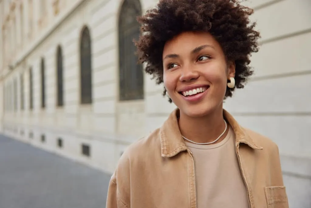 Smiling young woman with curly hair standing outdoors near a building with arched windows