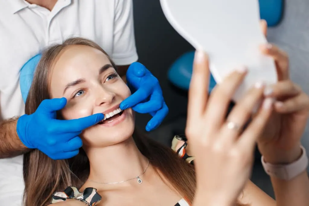 Happy woman in a dental chair holding a mirror while dentist checks her smile.